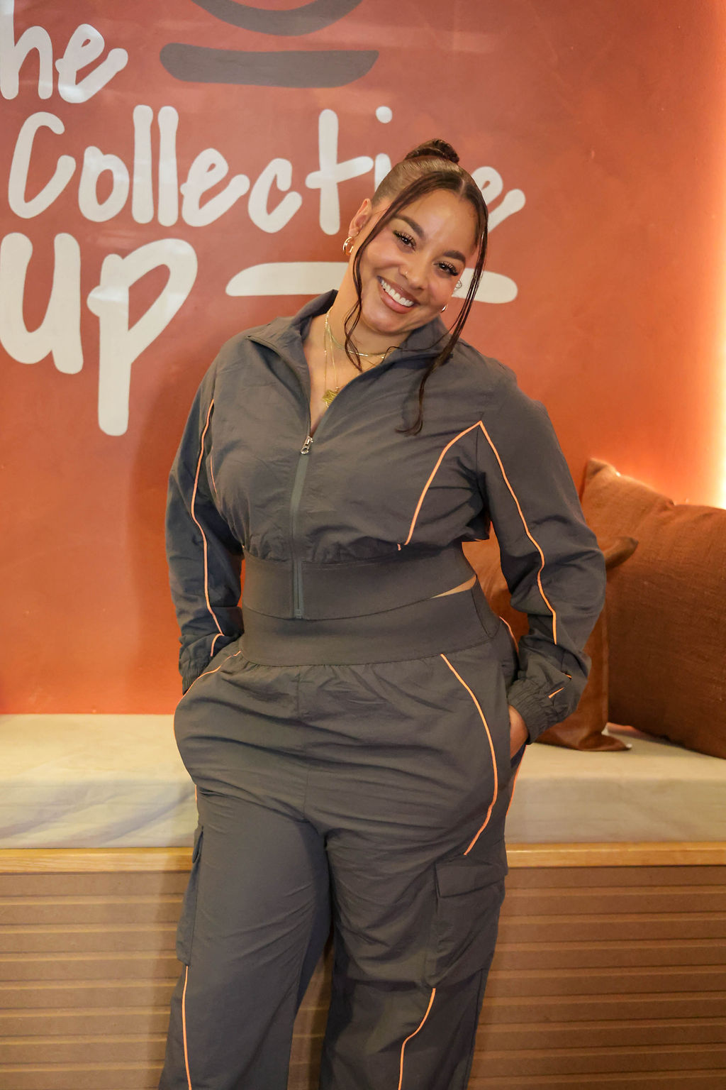 A smiling guest in a charcoal tracksuit with orange piping standing in front of the terracotta wall painted with The Collective Cup