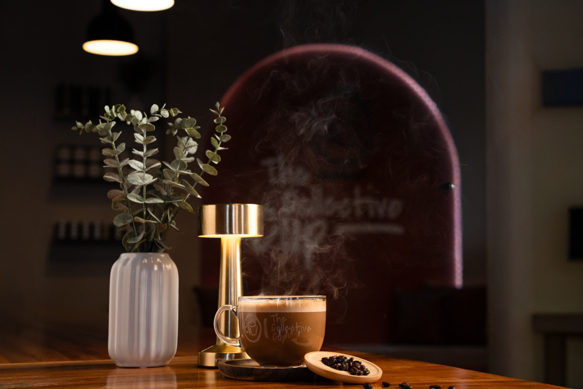 A steaming glass mug of coffee from The Collective Cup beside roasted beans and a warm brass lamp