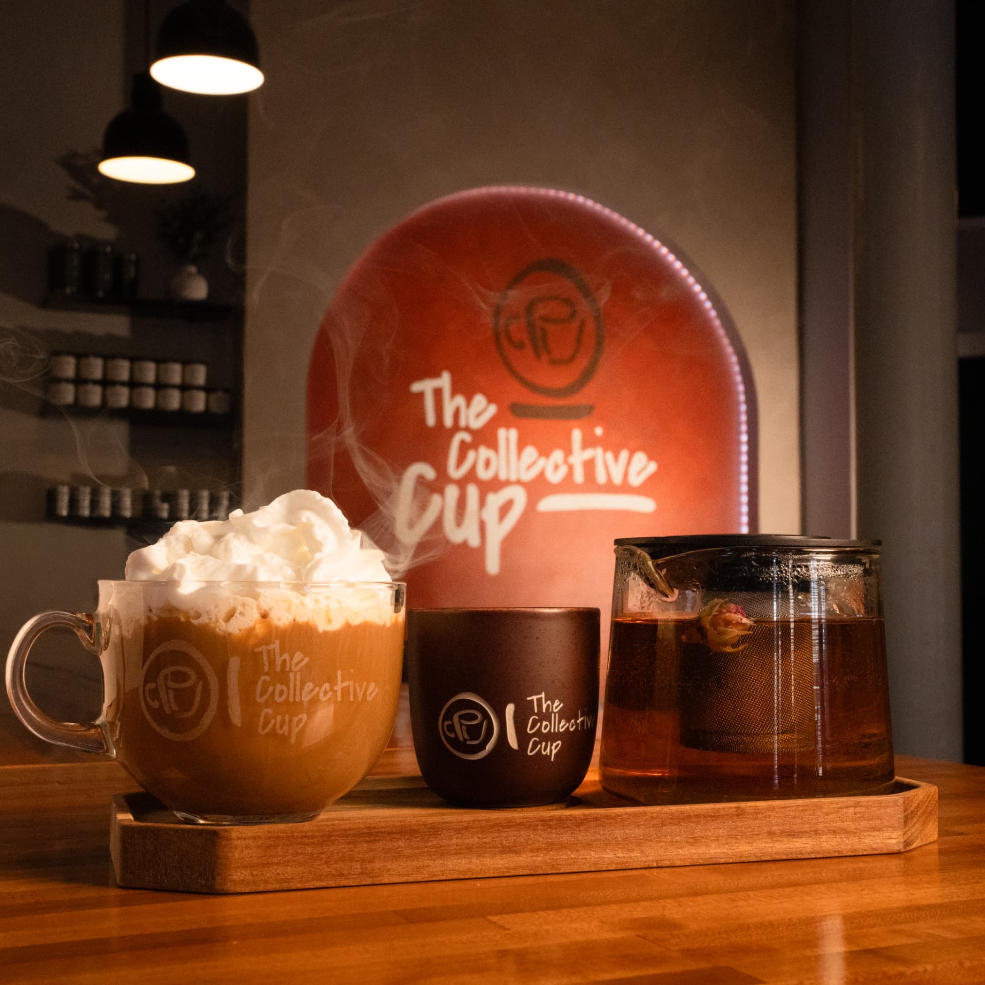 A wooden tray holding a whipped cream coffee, a branded ceramic cup and a glass teapot of tea